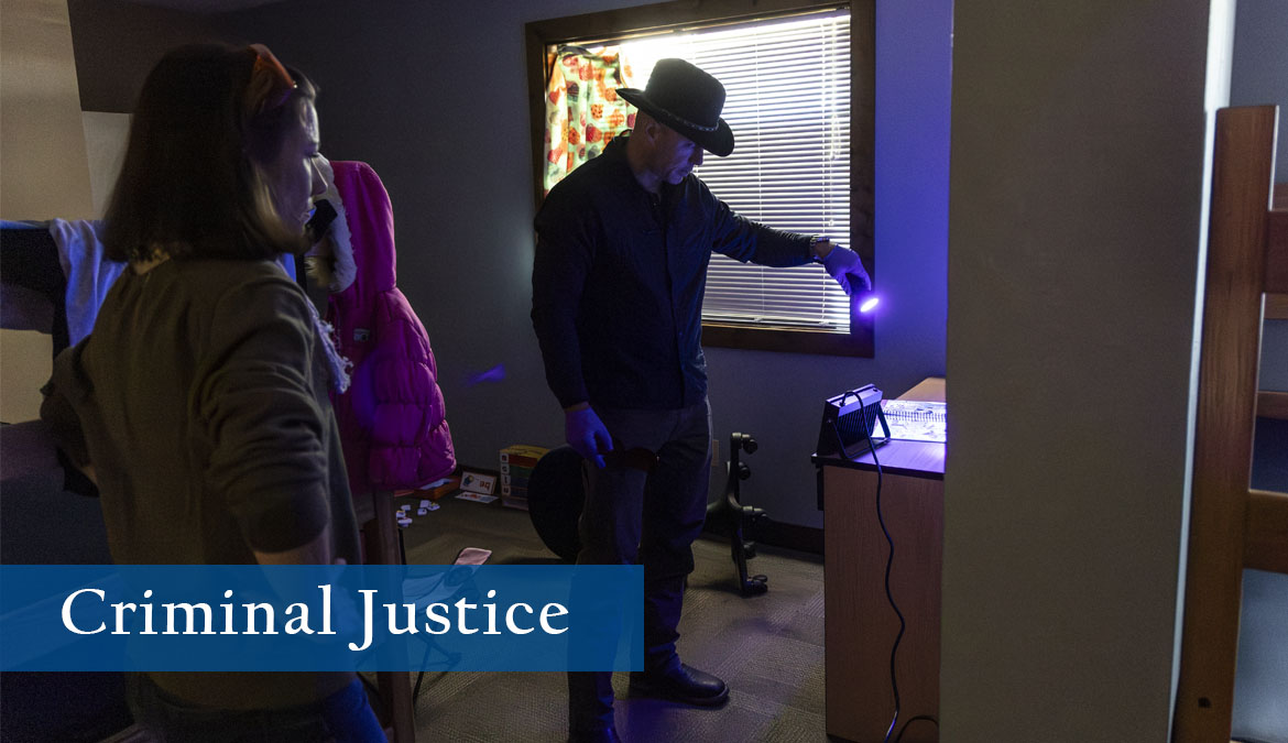 Criminal Justice Criminal Justice student shines a purple light to reveal evidence during a mock investigation of a crime scene in a bedroom setting with the blinds drawn.