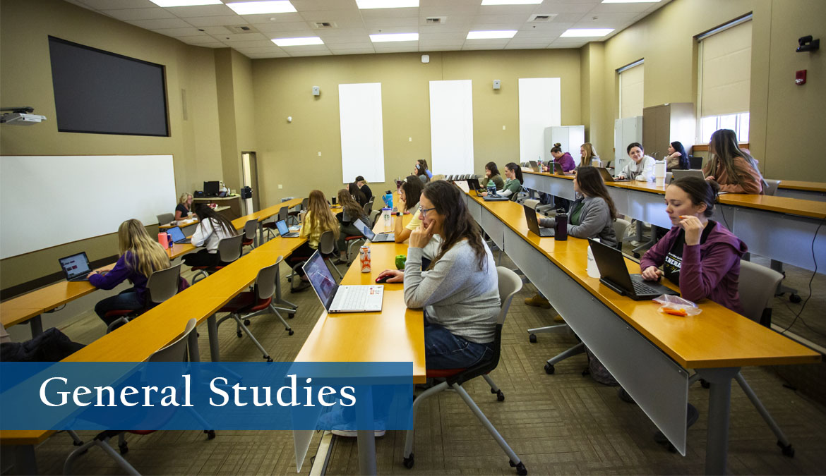 General Studies Photo of a tiered, brightly lit classroom with rows of students at their desks with laptops.