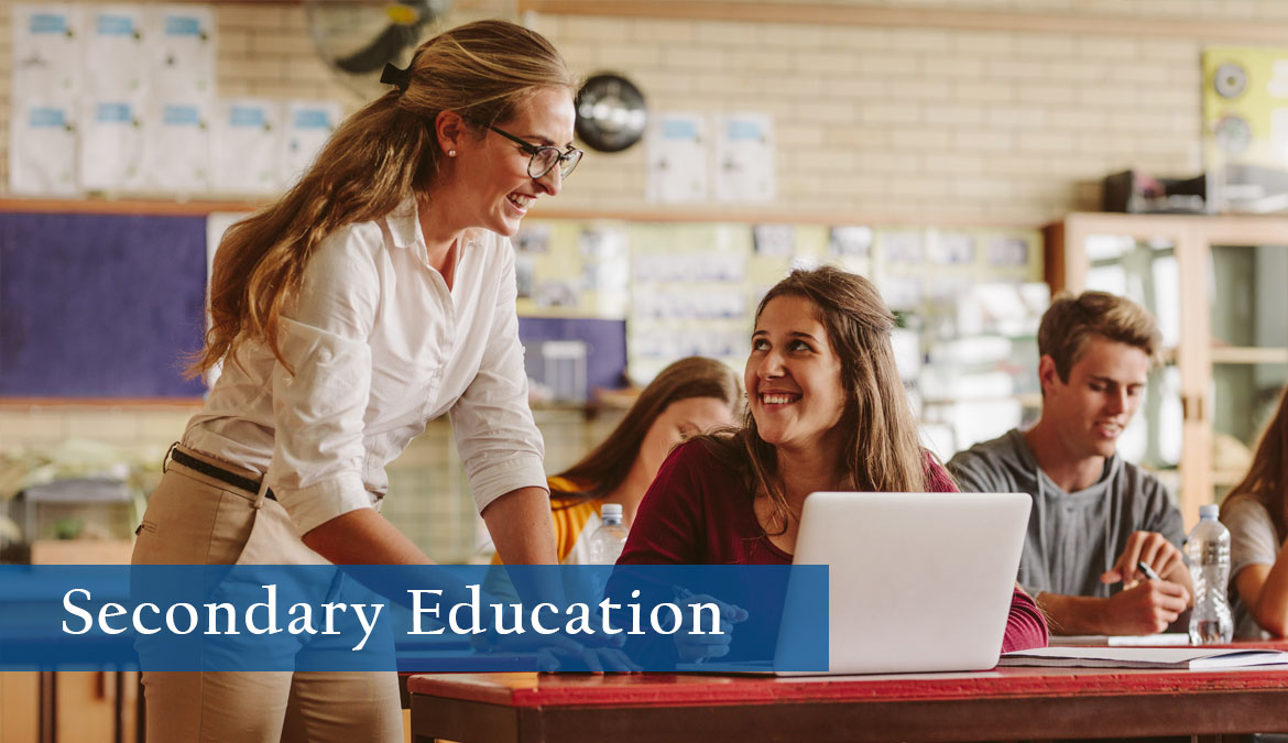 Secondary Education Photo of teacher working with a high school student who is at their desk with a laptop in a classroom.