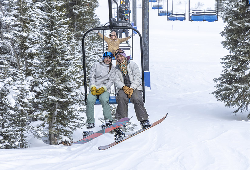 Snowboarding at Antelope Butte Ski Area snowboarding at antelope butte, two female students smiling and riding the chairlift with one behind them also smiling and putting her hands up in the air excited